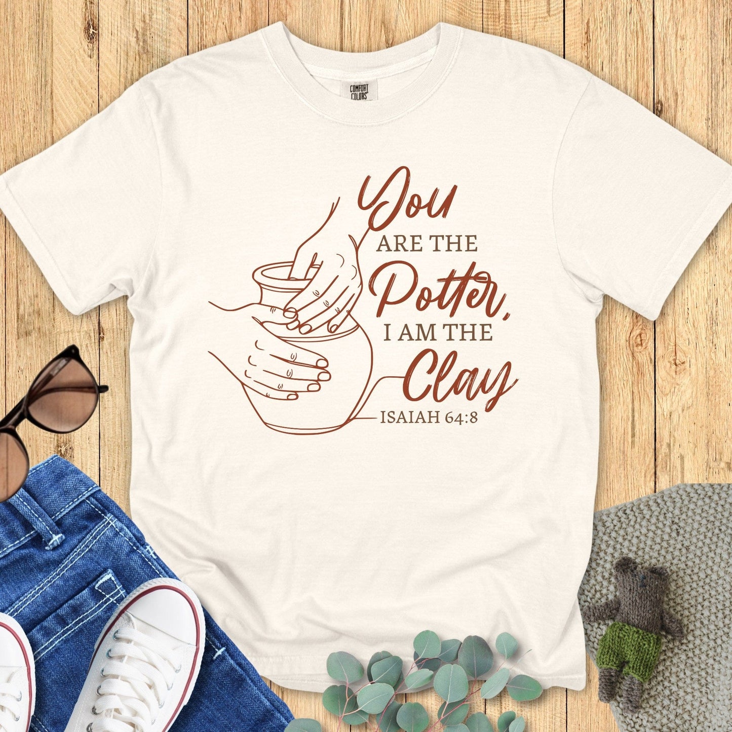 Comfort Colors Christian t-shirt in Ivory with Isaiah 64:8 “You are the Potter, I am the Clay” scripture and pottery hands line art. Faith-based shirt styled in a flat lay with denim, white sneakers, sunglasses, and cozy knit blanket.