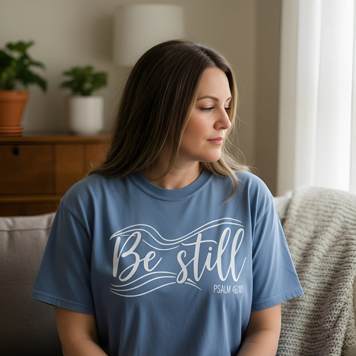 Woman wearing a “Be Still” Christian t-shirt in a quiet indoor setting, representing encouragement and scripture-based faith tees.