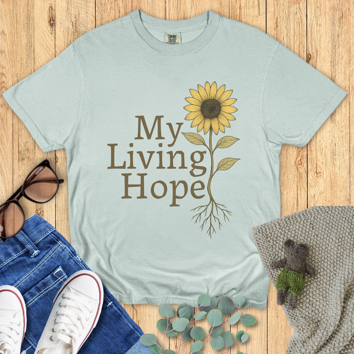 Light blue t-shirt with sunflower graphic and 'My Living Hope' text on a wooden surface with accessories.
