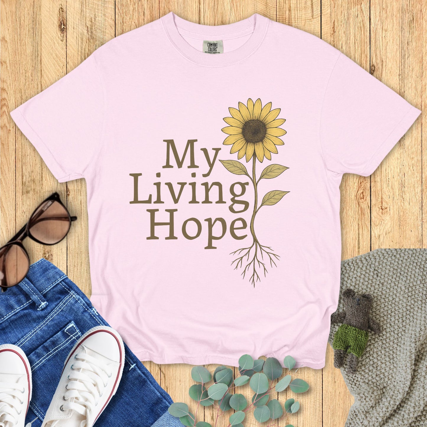 Pink t-shirt with sunflower graphic and 'My Living Hope' text on a wooden surface with accessories.