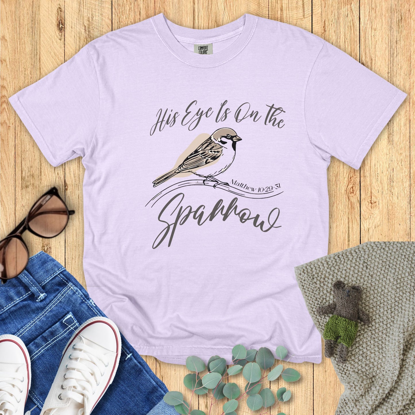 Light purple t-shirt with bird graphic and text on a wooden surface with accessories.