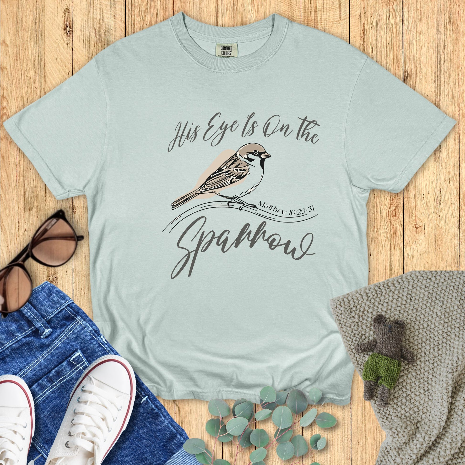 Light blue t-shirt with a bird graphic and text on a wooden surface with accessories.