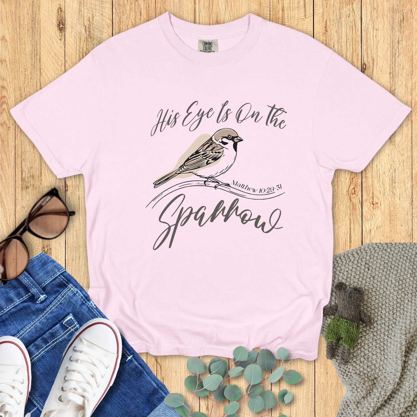 Pink t-shirt with bird graphic and text on a wooden surface with accessories.