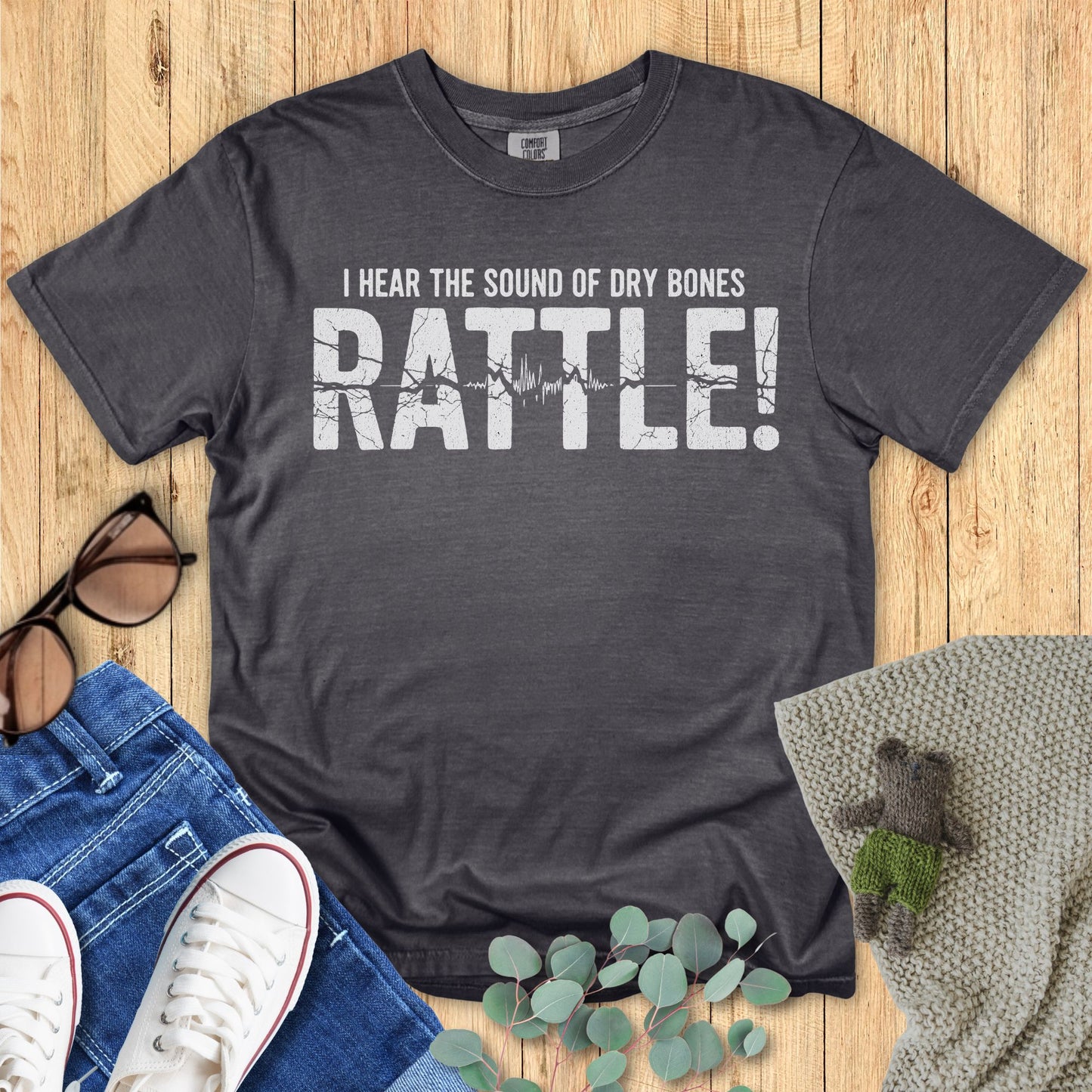Dark gray t-shirt with 'I hear the sound of dry bones RATTLE!' text on a wooden surface with accessories.