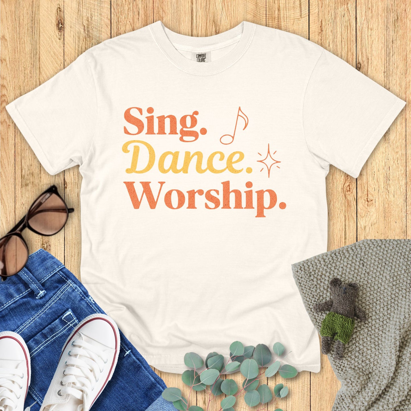 Ivory t-shirt with the words “Sing. Dance. Worship.” in orange and yellow with playful music note and sparkle graphics.
