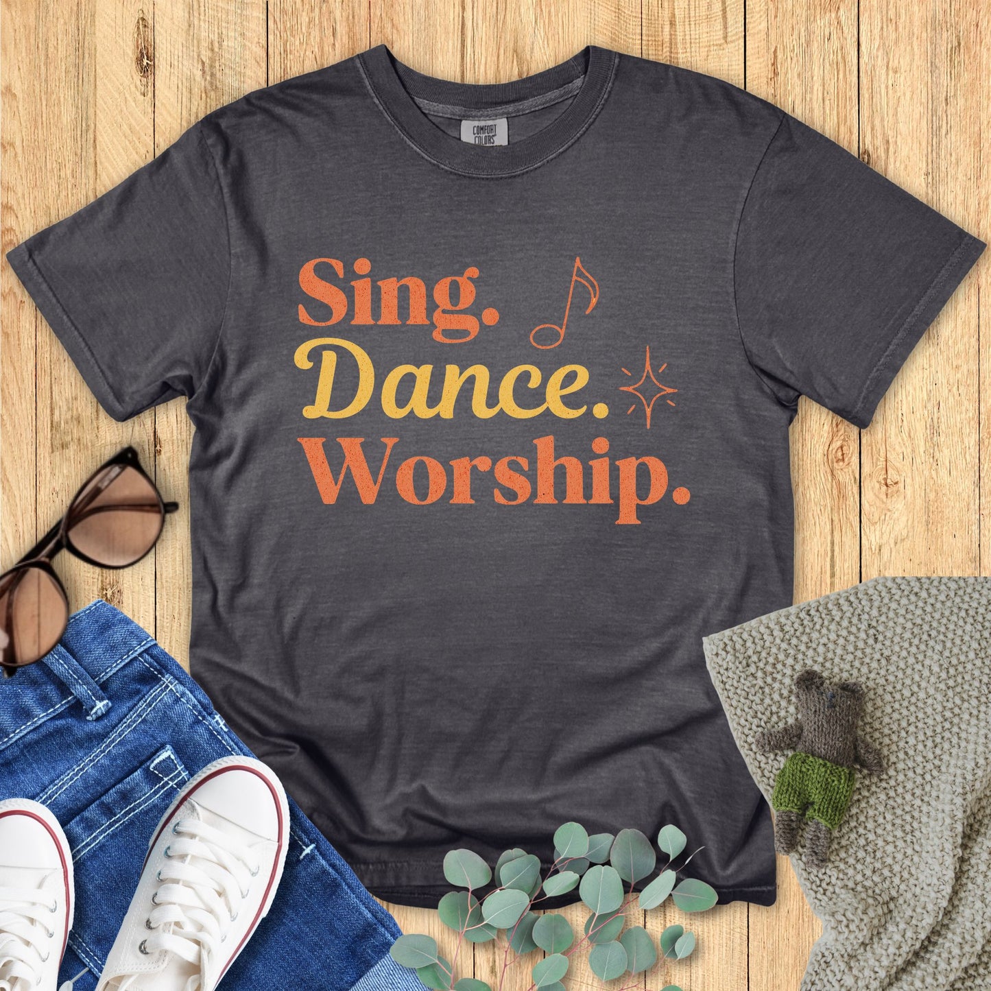 Gray t-shirt with 'Sing. Dance. Worship.' text on a wooden surface with accessories.