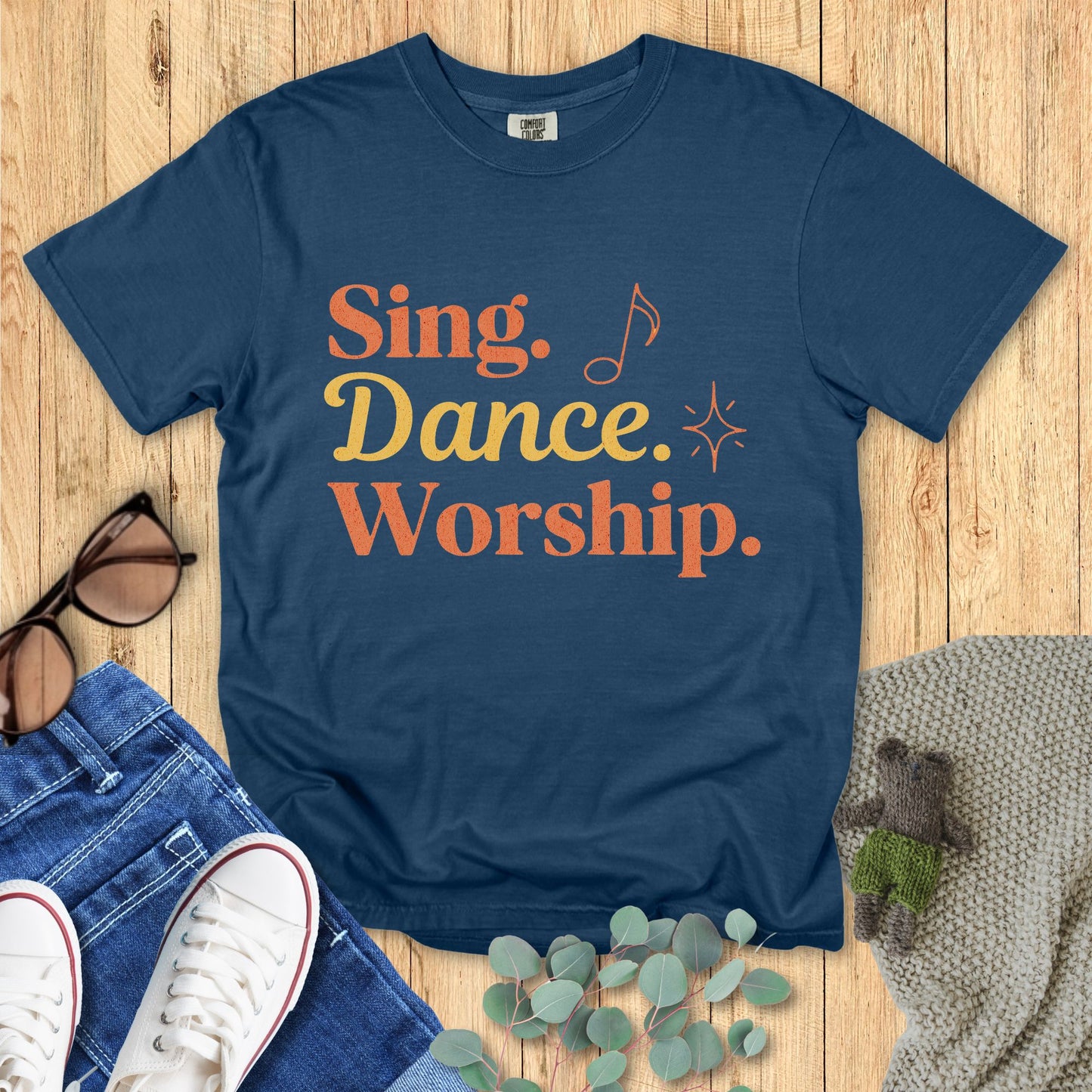 Blue t-shirt with 'Sing. Dance. Worship.' text on a wooden surface with accessories.