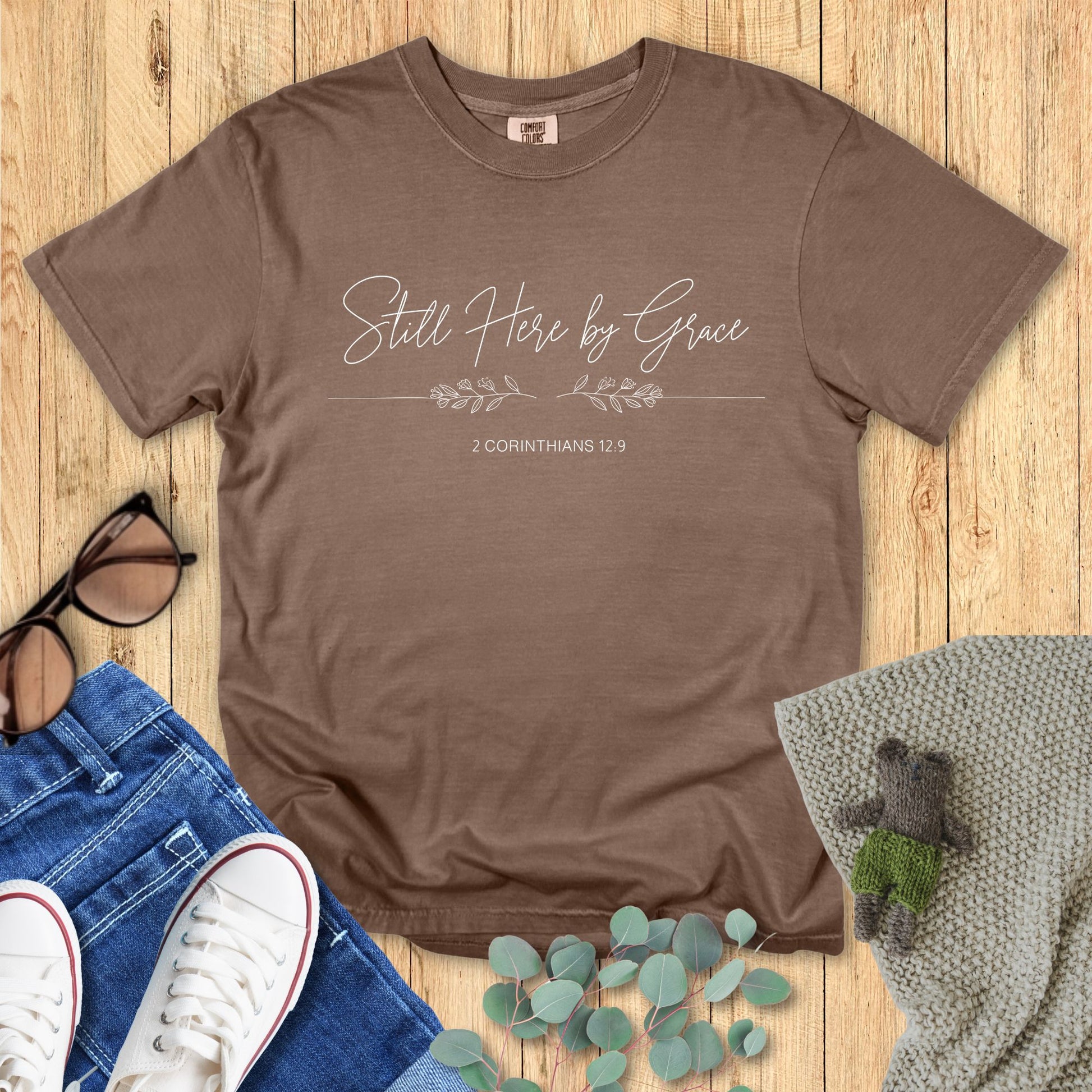 Brown t-shirt with 'Still Here by Grace' text on a wooden surface with accessories.