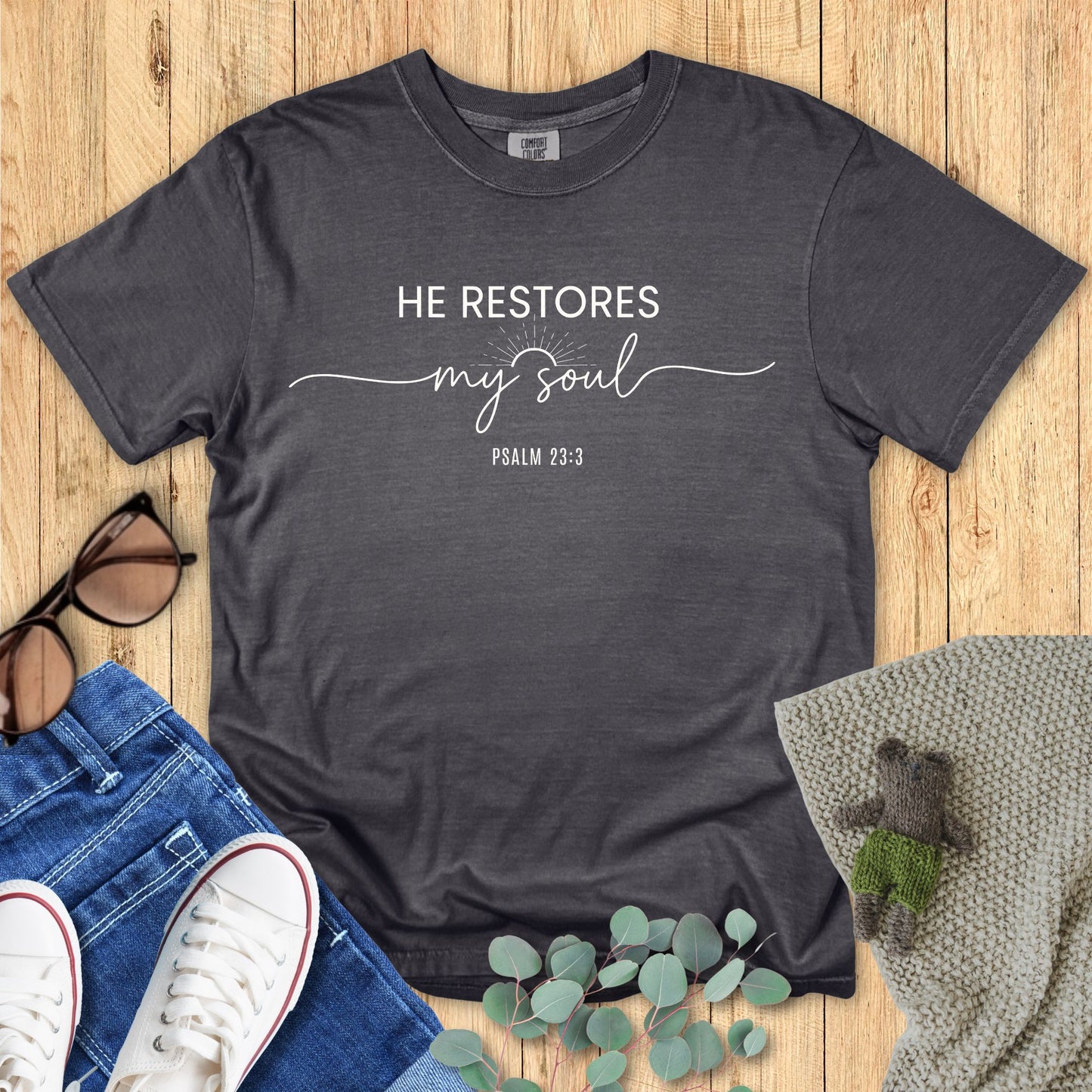 Dark gray t-shirt with 'He restores my soul' text on a wooden surface with accessories.
