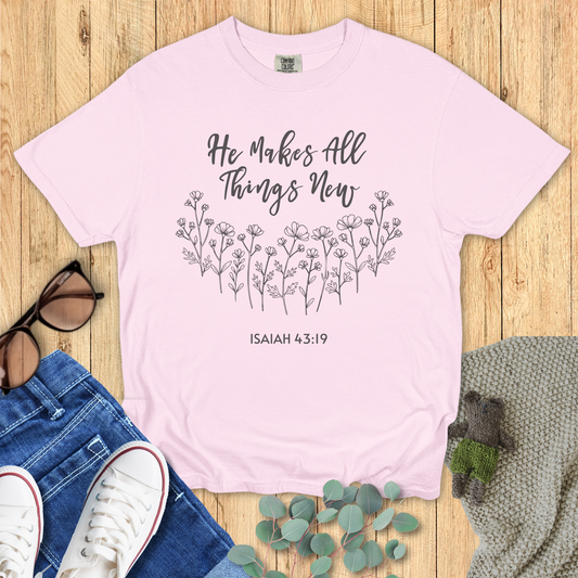 Pink t-shirt with 'He Makes All Things New' text on a wooden surface with jeans and sneakers.