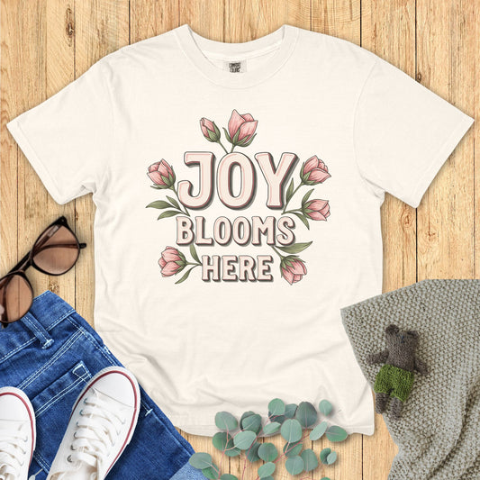 Ivory White t-shirt with 'Joy Blooms Here' design on a wooden surface with accessories.