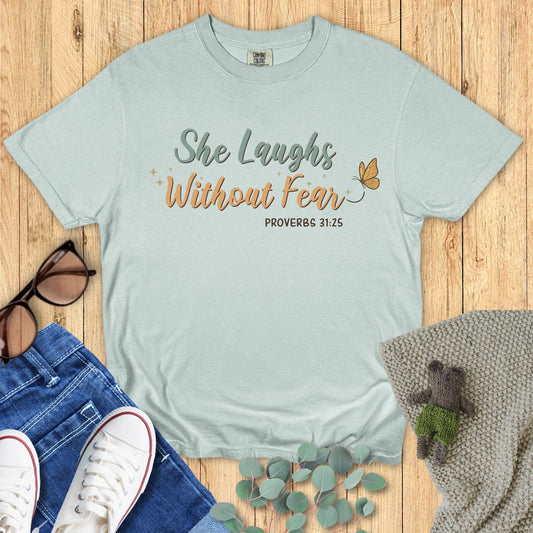 Light blue t-shirt with 'She Laughs Without Fear' text on a wooden surface with accessories.