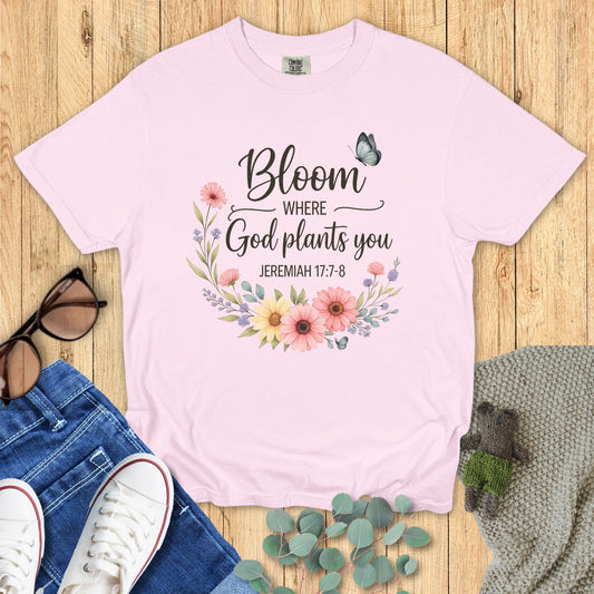 Pink t-shirt with floral design and text on a wooden surface with accessories.