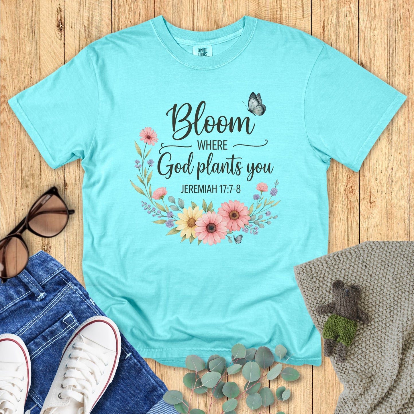 Light blue t-shirt with floral design and text on a wooden surface with accessories.