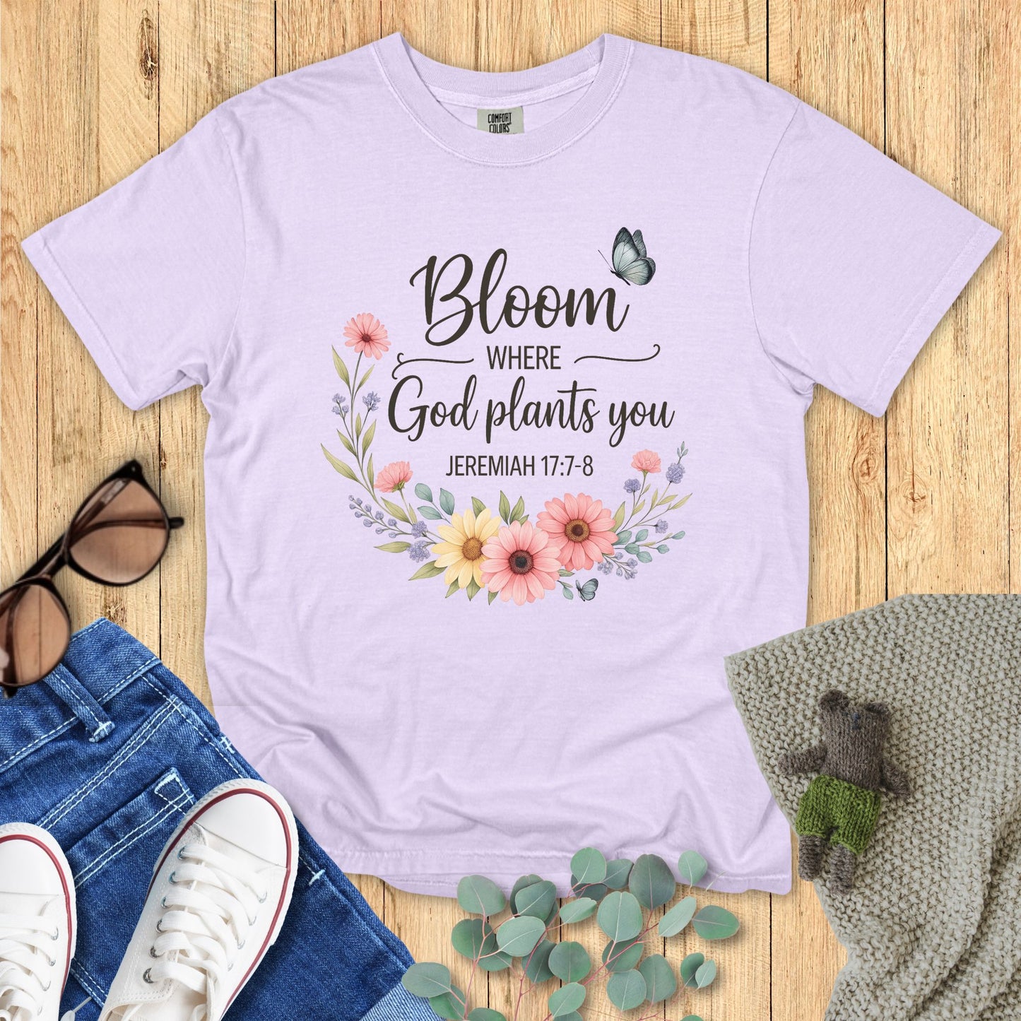 Lavender t-shirt with floral design and text on a wooden surface with accessories.