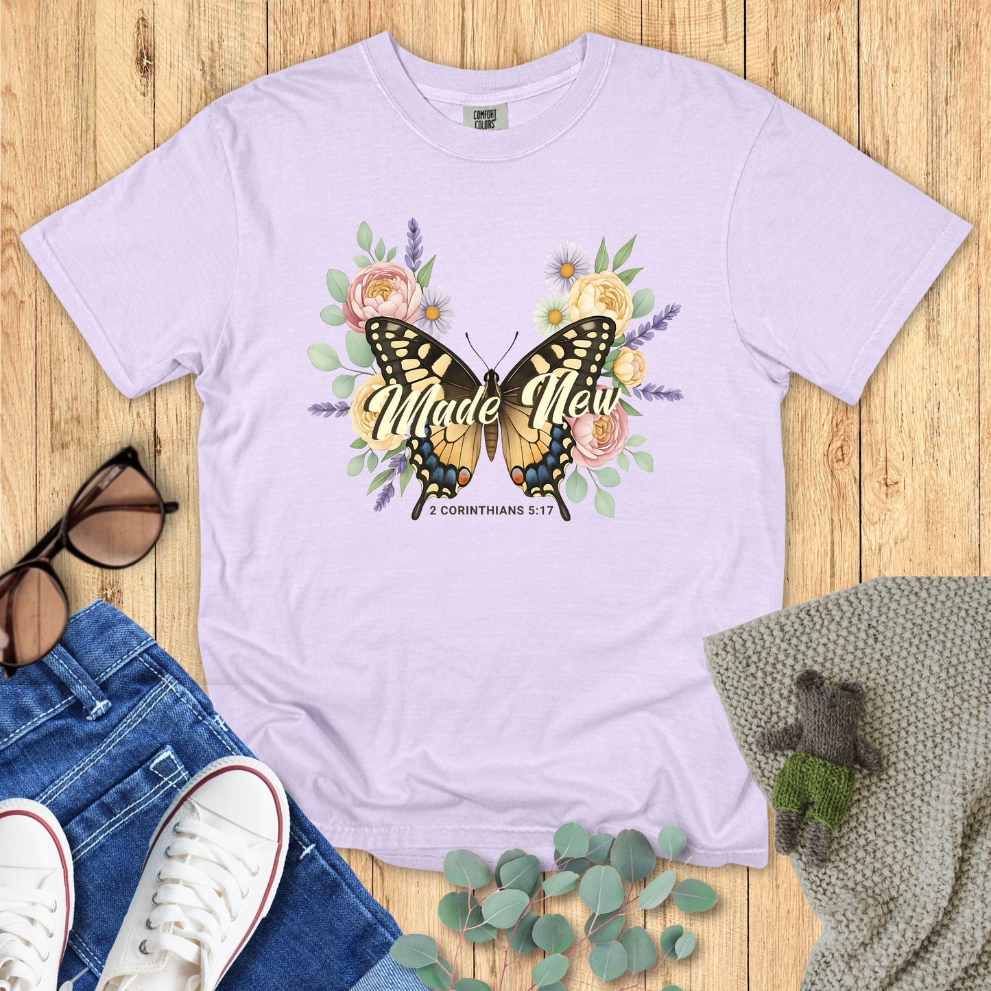 Lavender t-shirt with butterfly graphic and 'Made New' text on a wooden surface with accessories.
