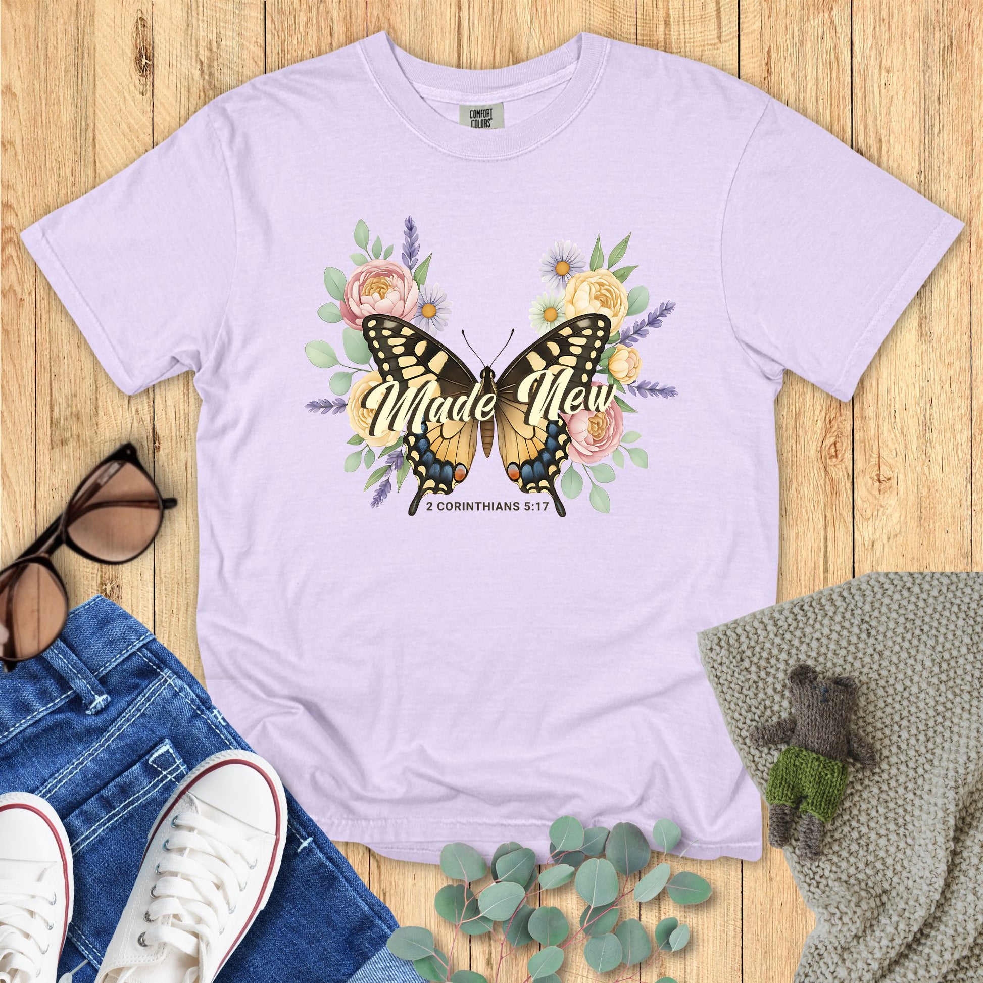Lavender t-shirt with butterfly graphic and 'Made New' text on a wooden surface with accessories.