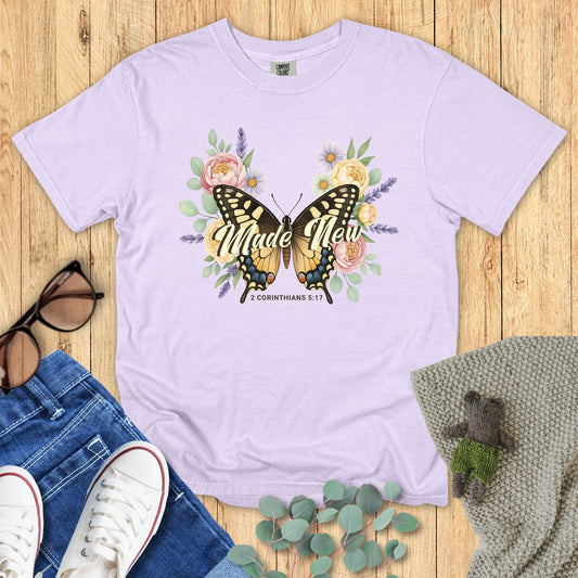 Lavender t-shirt with butterfly graphic and 'Made New' text on a wooden surface with accessories.