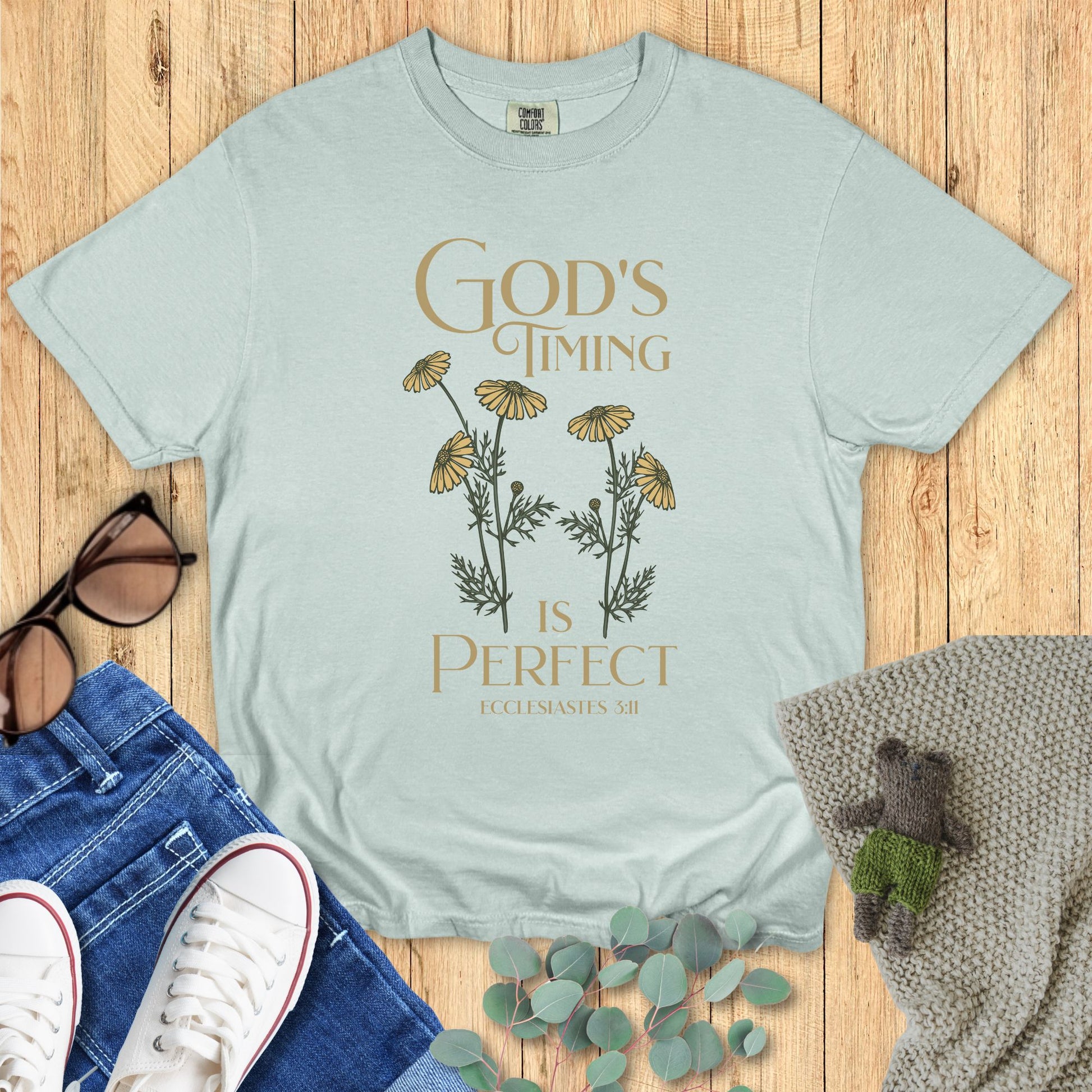 Light blue t-shirt with floral design and text on a wooden surface with accessories.