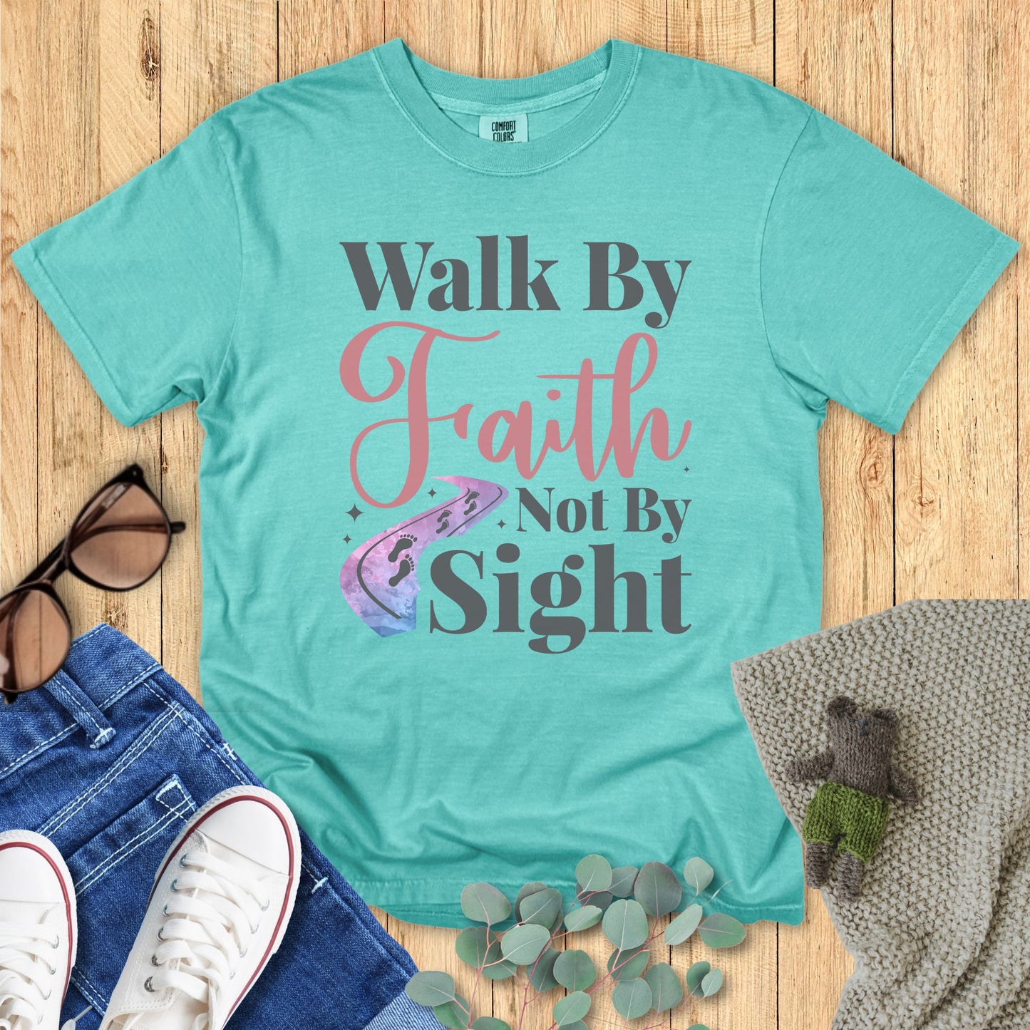 Teal t-shirt with 'Walk By Faith Not By Sight' text on a wooden surface with accessories.