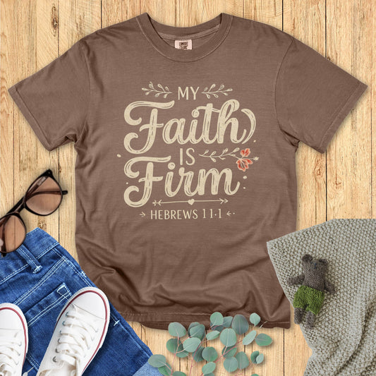 Brown t-shirt with 'My Faith Is Firm' text on a wooden surface with accessories.