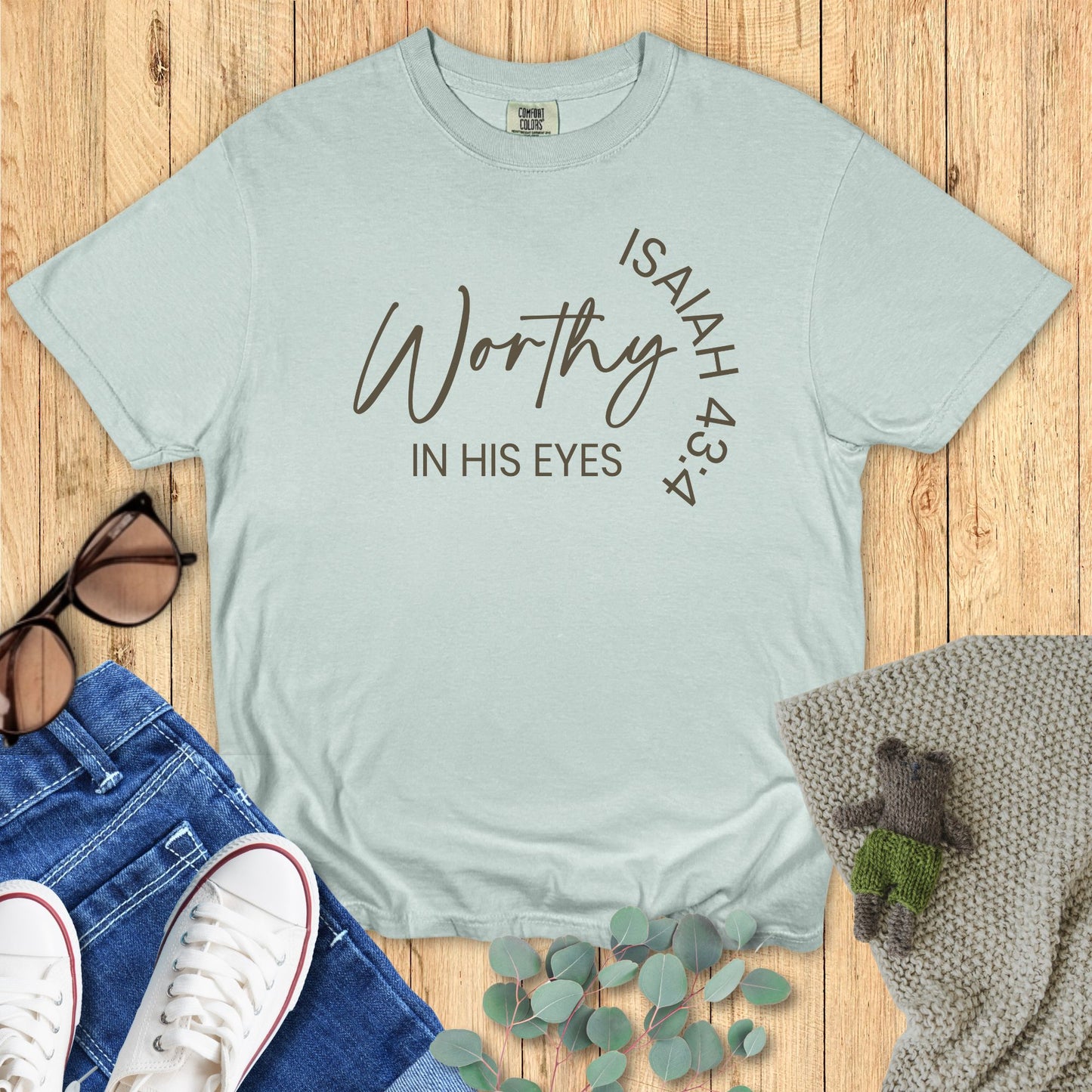 Light blue t-shirt with 'Worthy in His Eyes' text on a wooden surface with accessories.