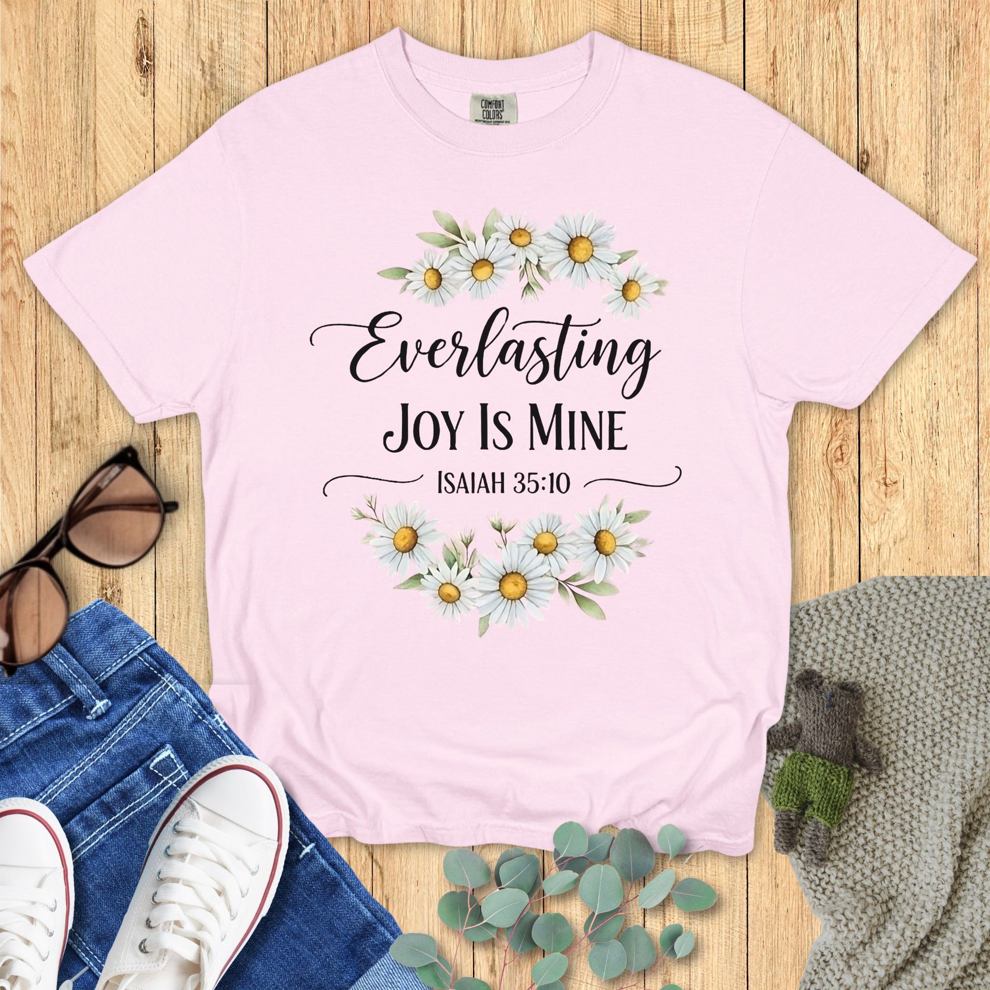 Pink t-shirt with floral design and text on a wooden surface with accessories.