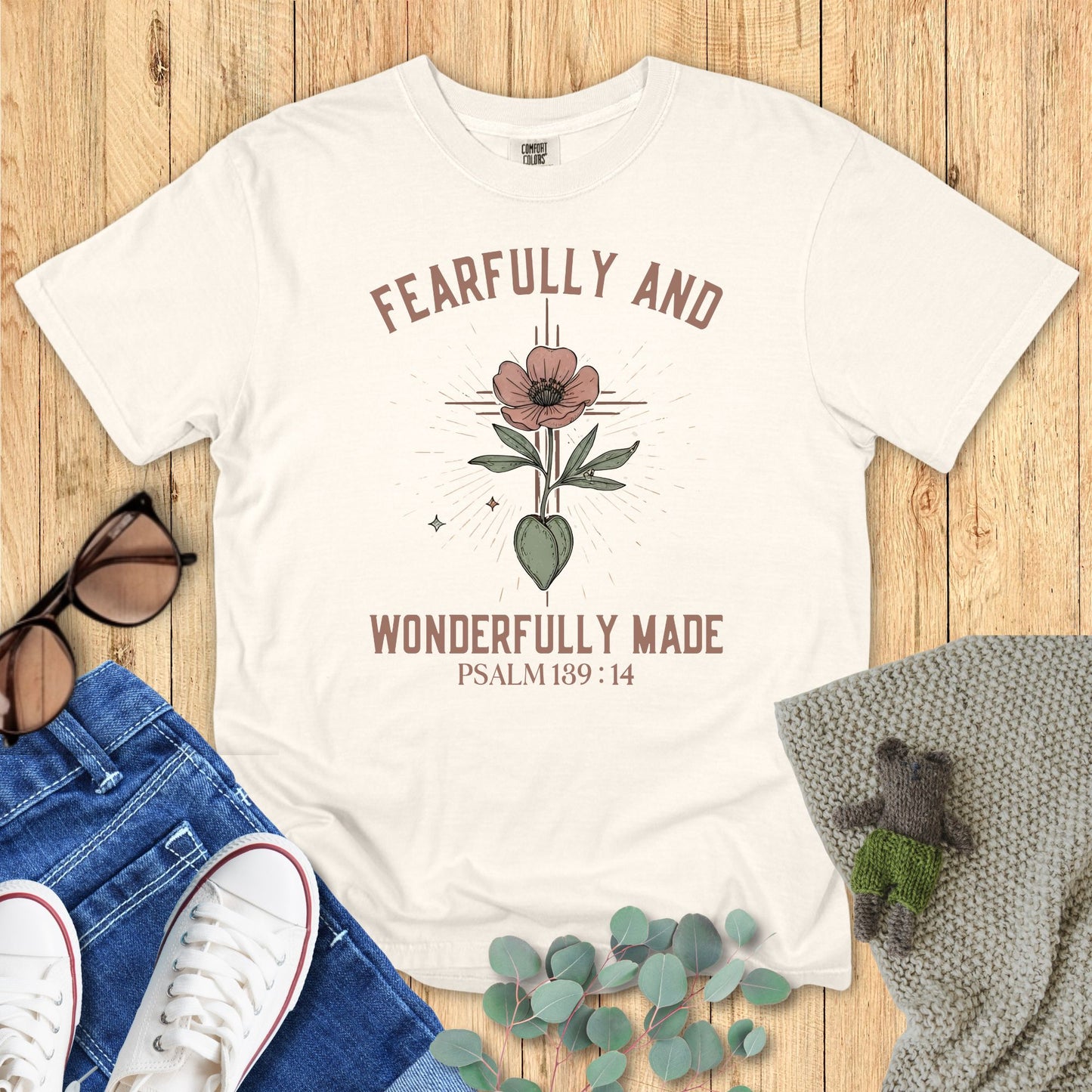 Ivory t-shirt with 'Fearfully and Wonderfully Made' design on a wooden surface with accessories.