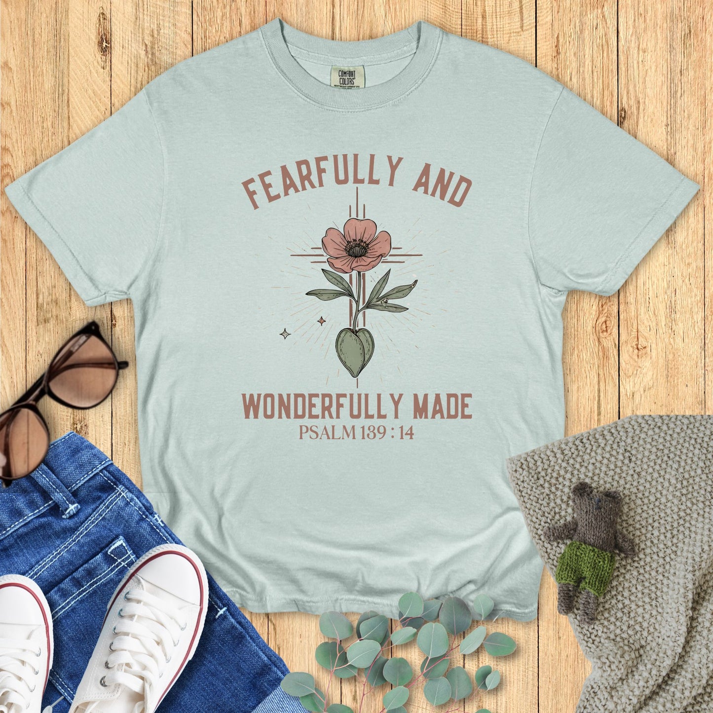 Light blue t-shirt with floral design and text on a wooden surface with accessories.