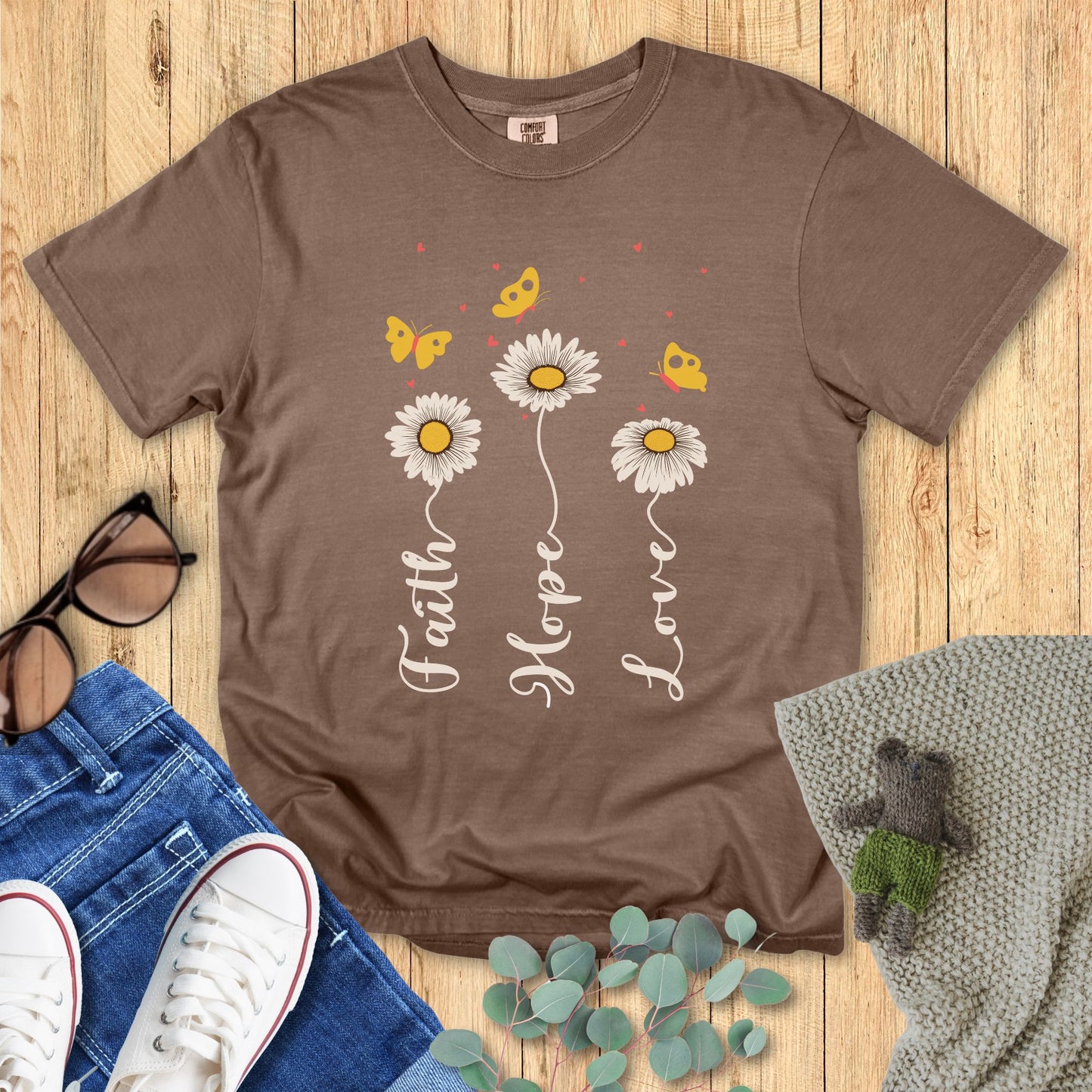 Brown t-shirt with floral design and text on a wooden surface with accessories.