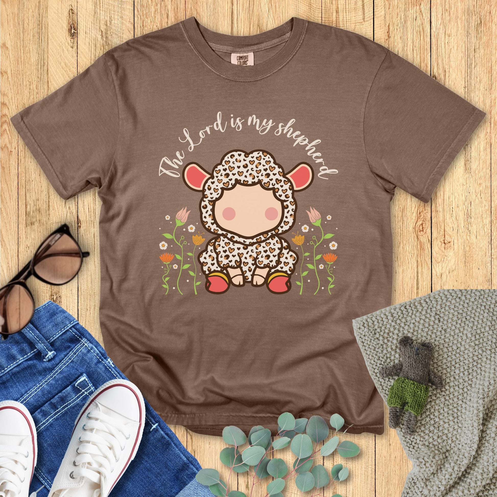 Brown t-shirt with a sheep graphic and 'The Lord is my shepherd' text on a wooden surface with accessories.