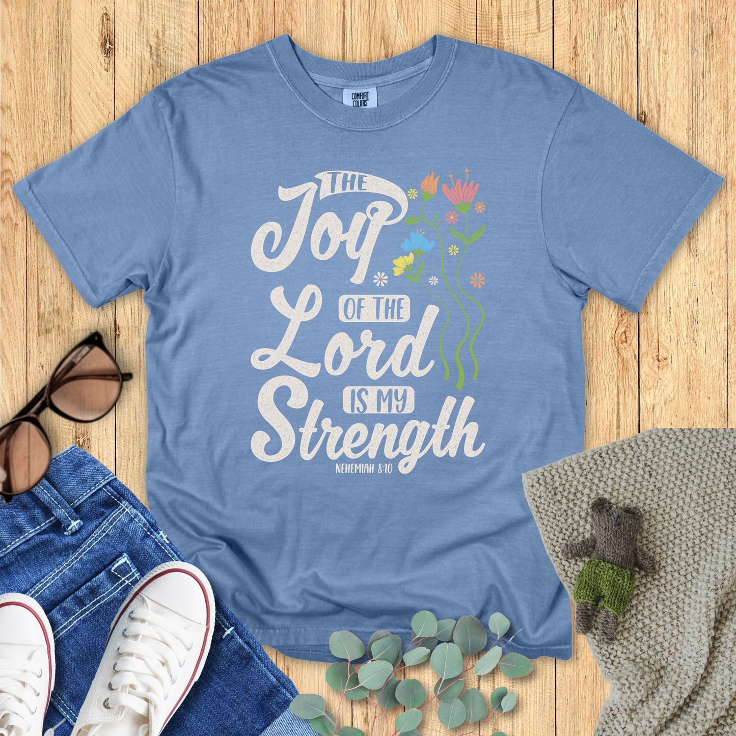 Blue t-shirt with 'The Joy of the Lord is My Strength' text on a wooden surface with accessories.