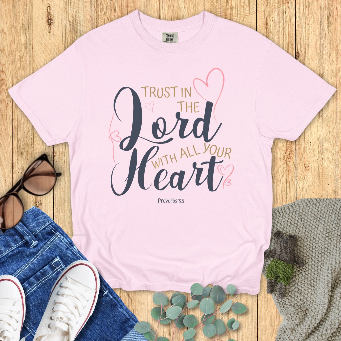 Pink t-shirt with 'Trust in the Lord with all your heart' text on a wooden surface with jeans and sneakers.