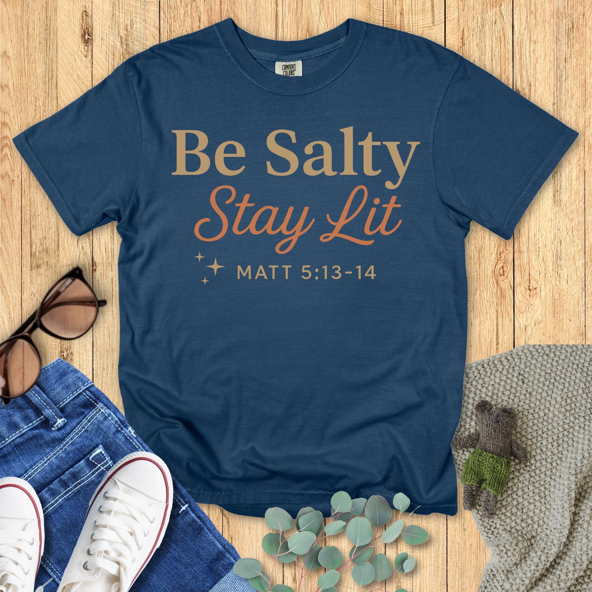 Blue t-shirt with 'Be Salty Stay Lit' text on a wooden surface with accessories.