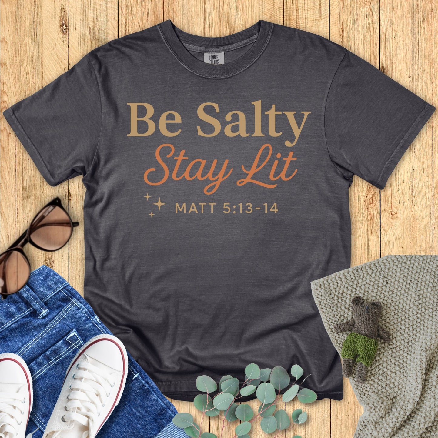 Gray t-shirt with 'Be Salty Stay Lit' text on a wooden surface with accessories.
