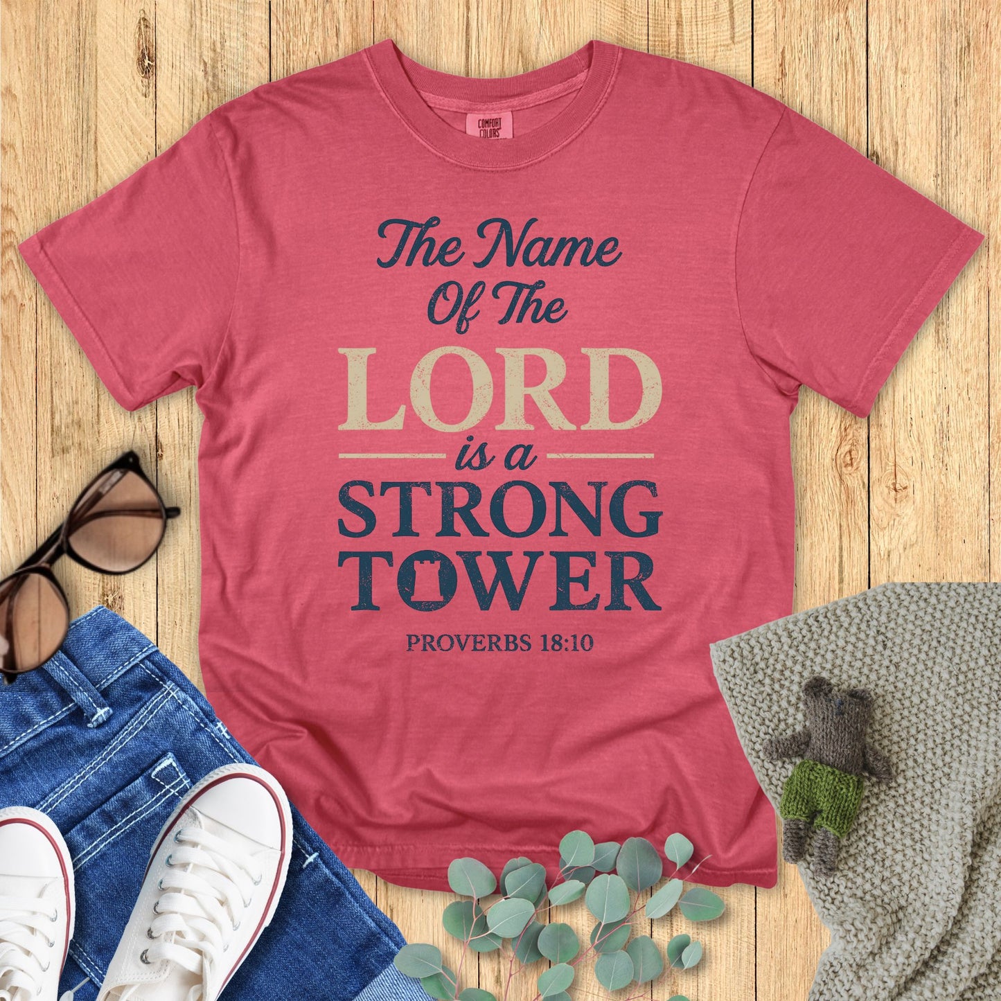 Red t-shirt with 'The Name of the Lord is a Strong Tower' text on a wooden surface with accessories.