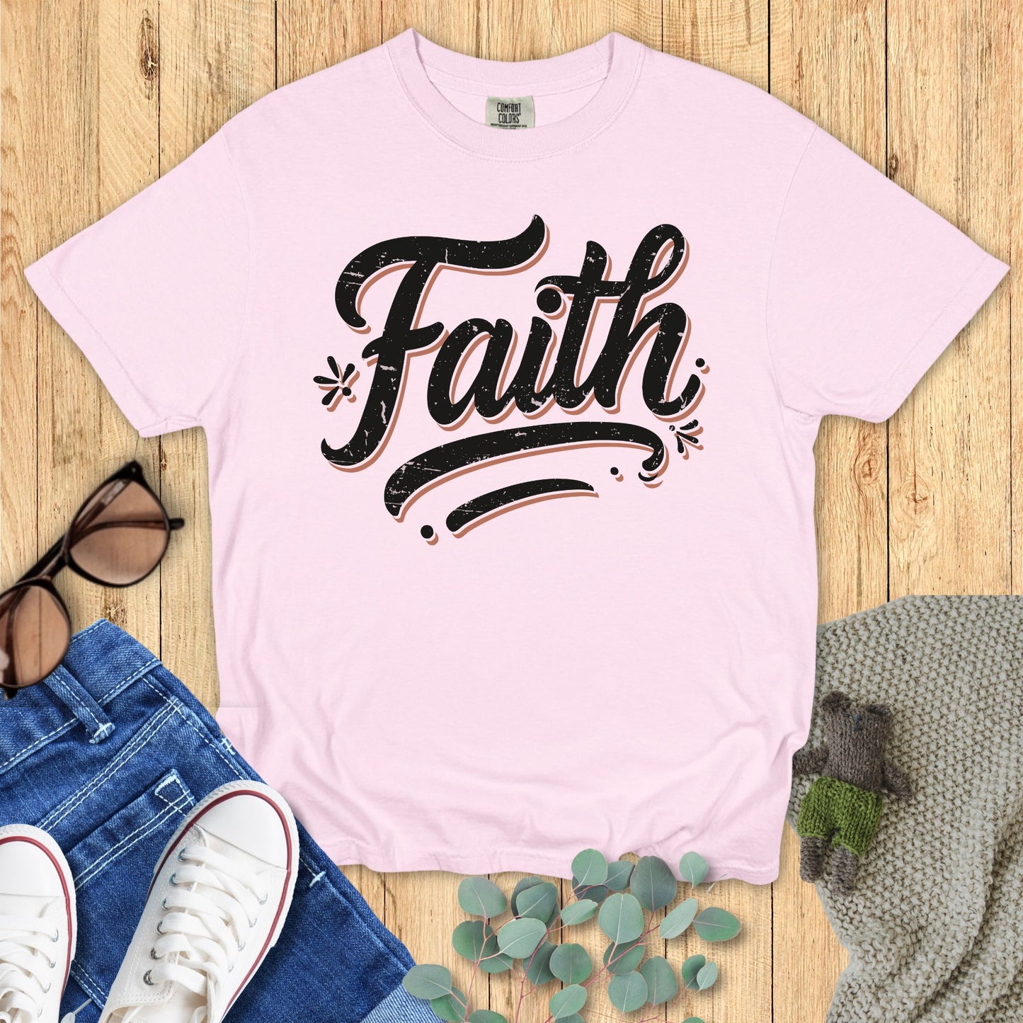Pink t-shirt with 'Faith' text on a wooden surface with jeans and sneakers.