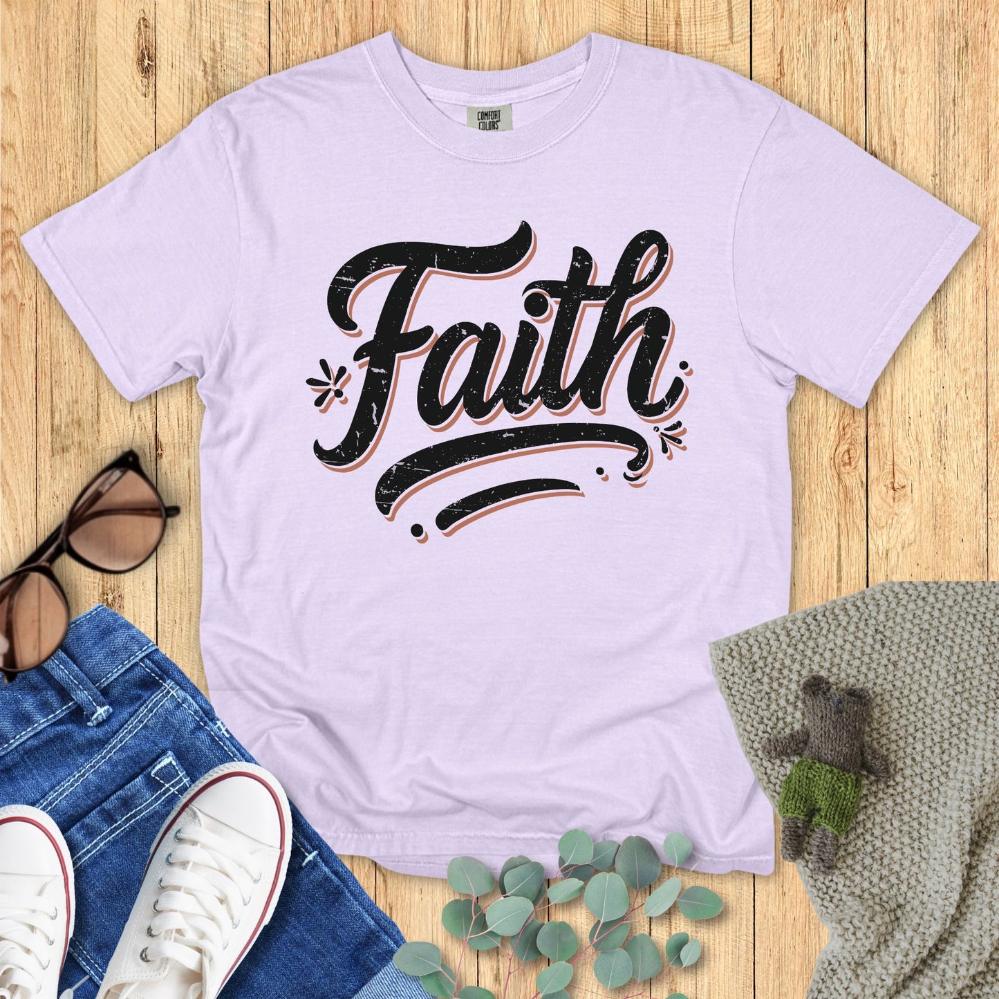 Light purple t-shirt with 'Faith' text on a wooden surface with accessories.