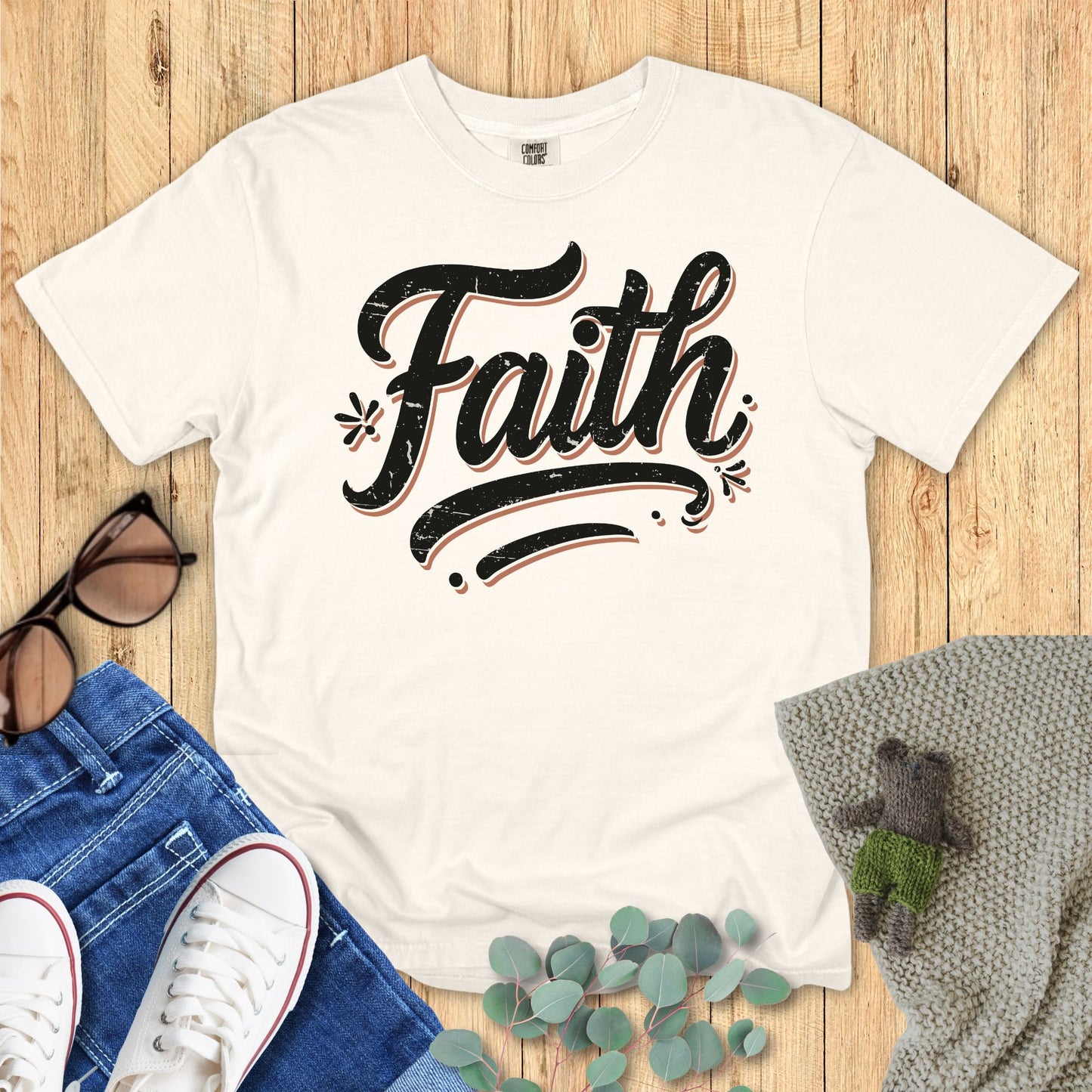 Ivory color t-shirt with 'Faith' text on a wooden surface with accessories.