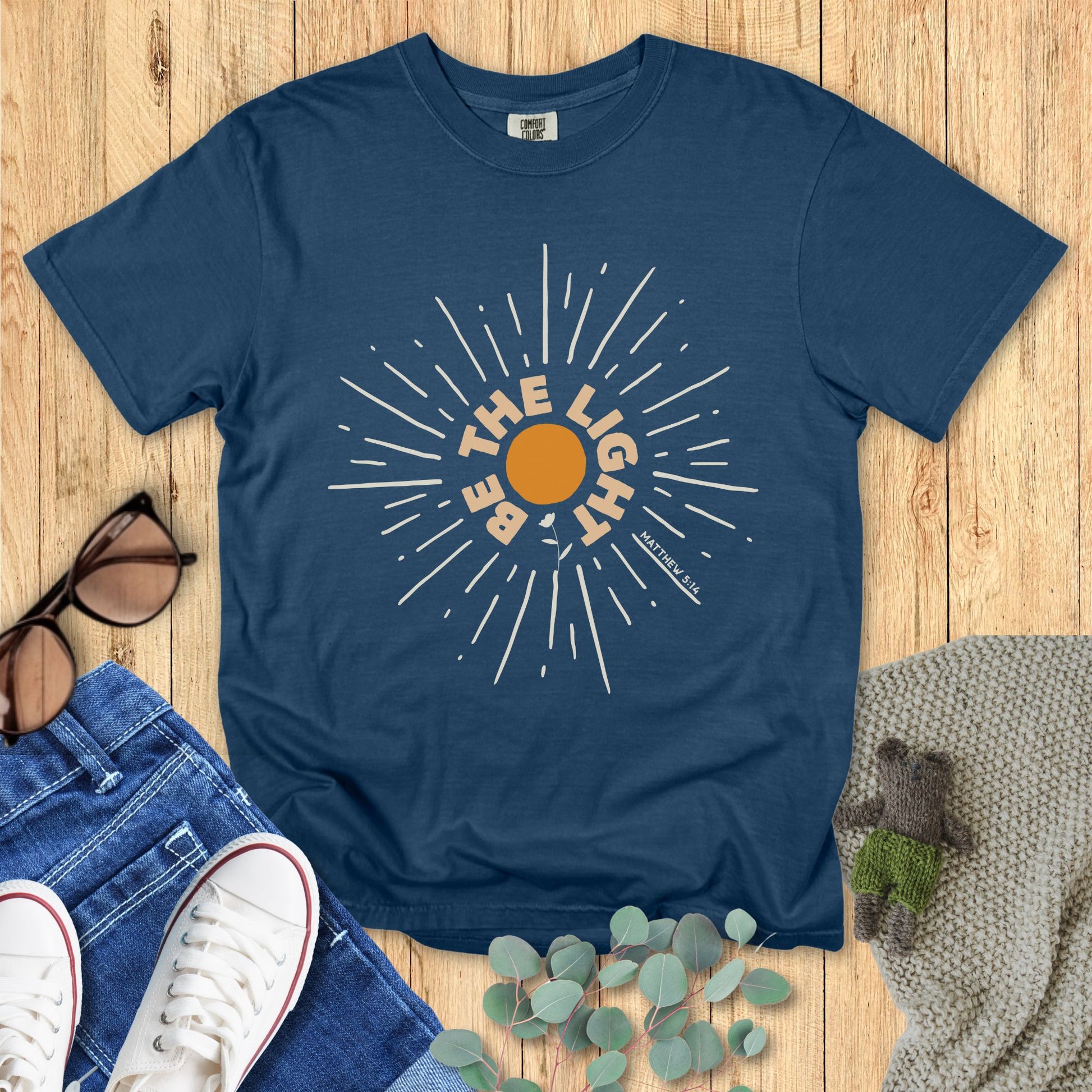 Blue t-shirt with sun graphic and text on a wooden surface with jeans and sneakers.