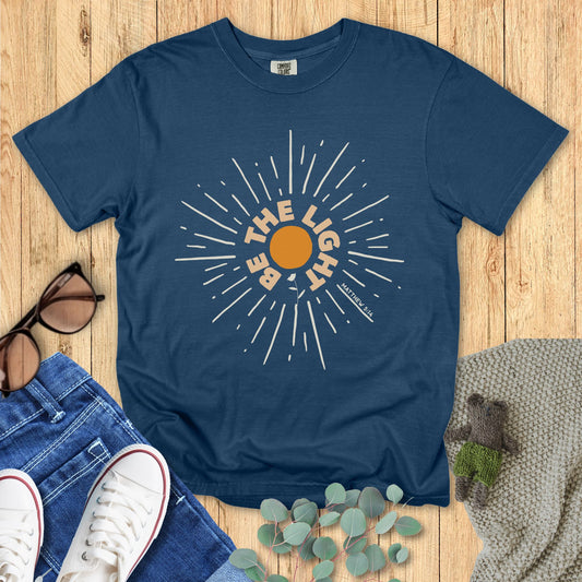 Blue t-shirt with sun graphic and text on a wooden surface with jeans and sneakers.
