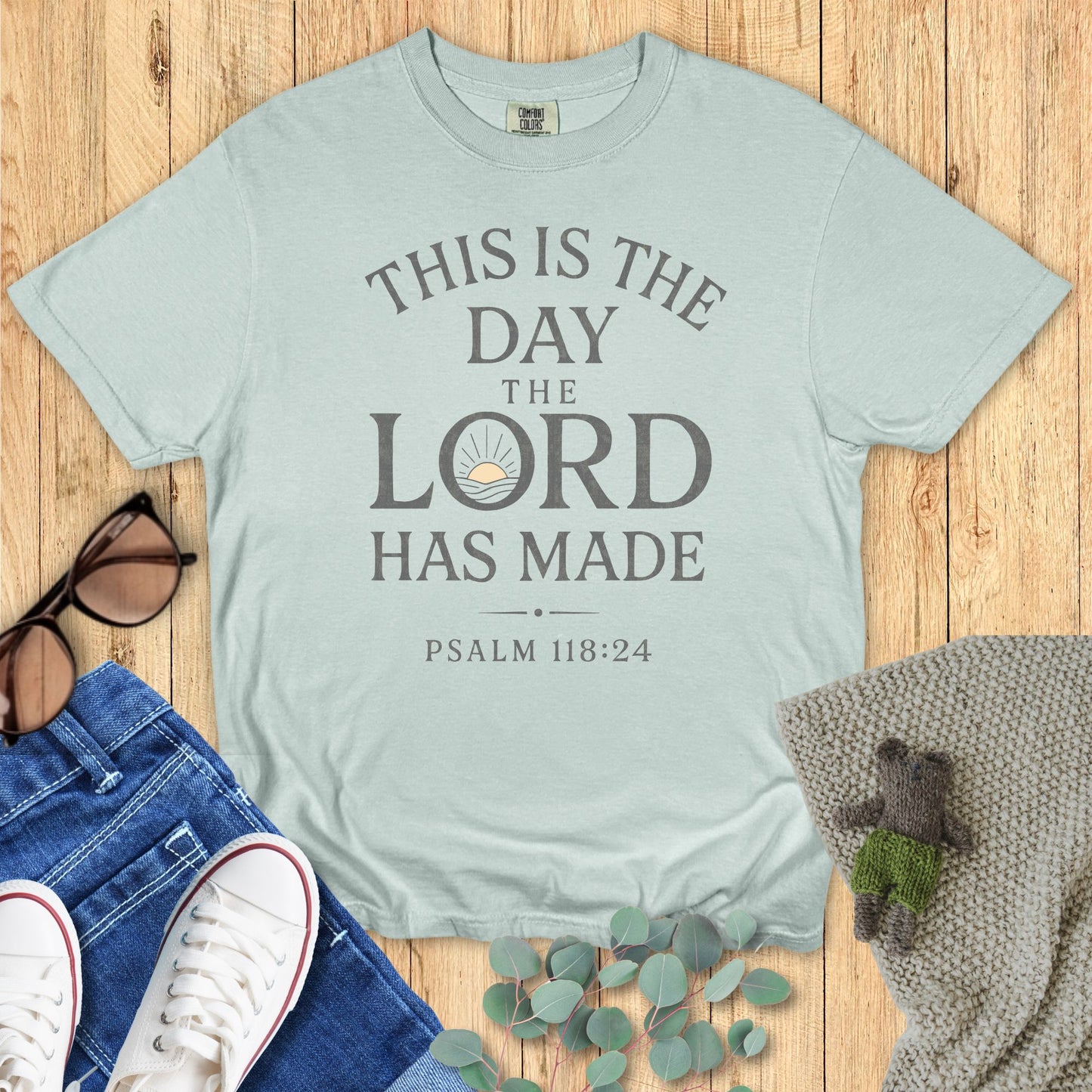 Light blue t-shirt with 'This is the day the Lord has made' text on a wooden surface with accessories.