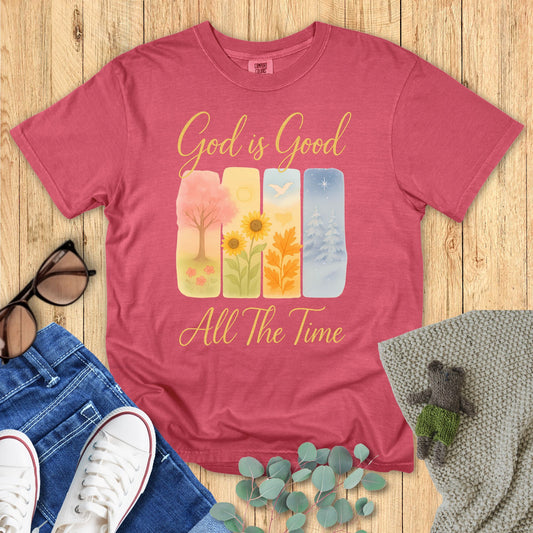 red t-shirt with 'God is Good All The Time' text and nature-themed design on a wooden surface with accessories.