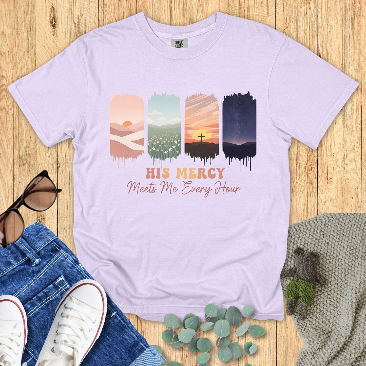 Lavender t-shirt with sunset design and text 'His Mercy Meets Me Every Hour' on a wooden surface with jeans and sneakers.