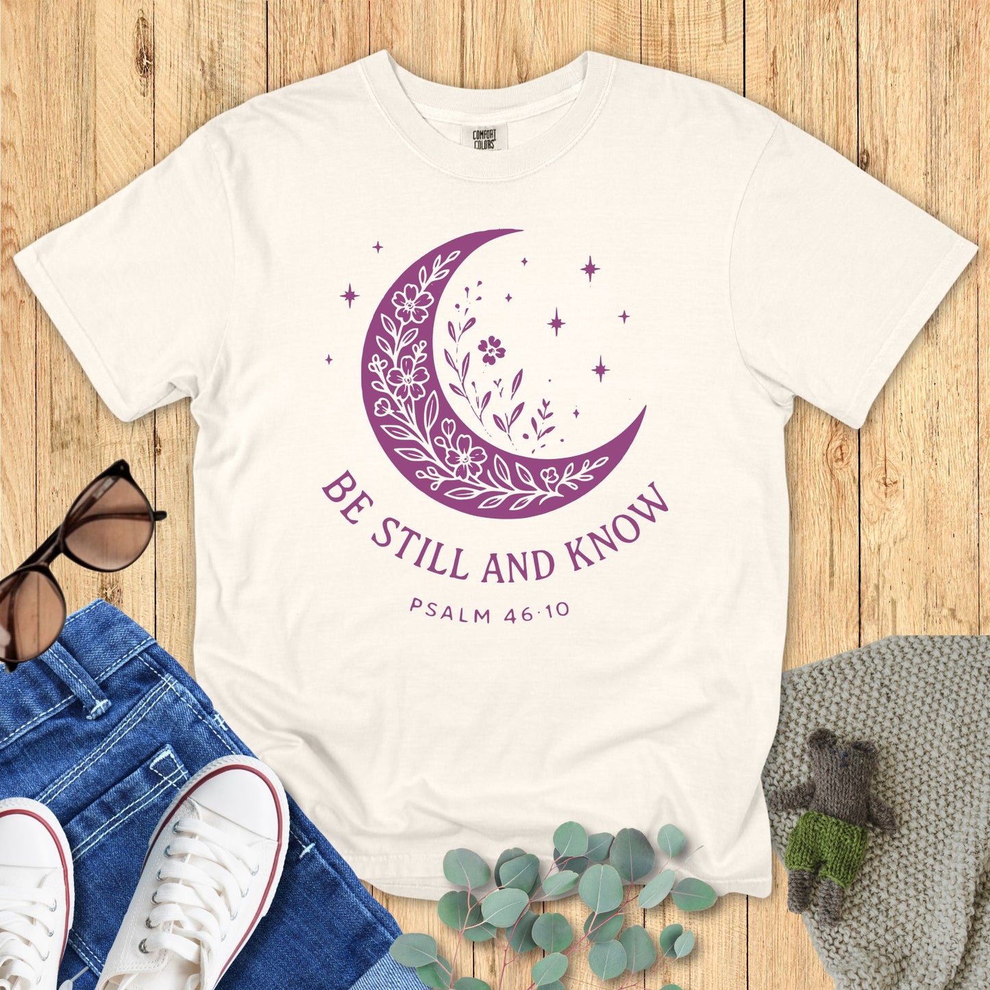 White t-shirt with purple moon design and 'Be Still and Know' text on a wooden surface with accessories.