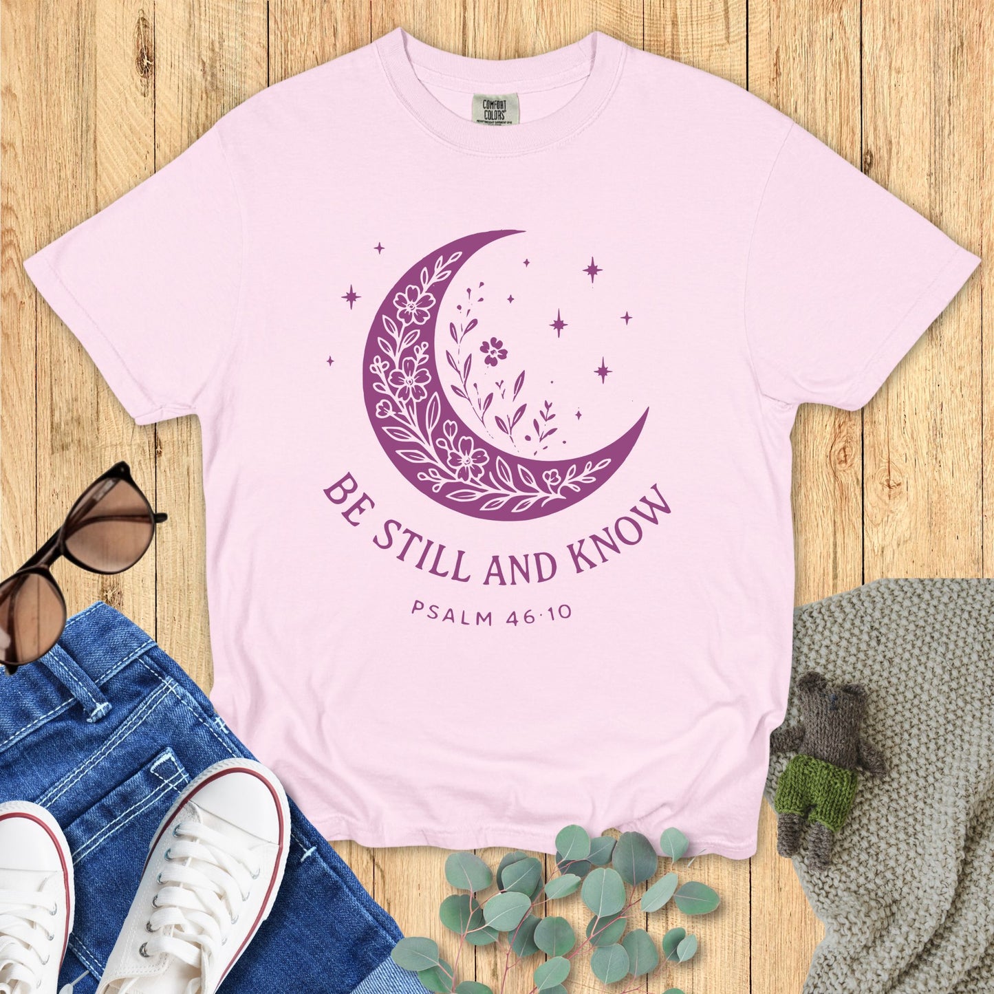 Pink t-shirt with purple moon design and 'Be Still and Know' text on a wooden surface with jeans and sneakers.
