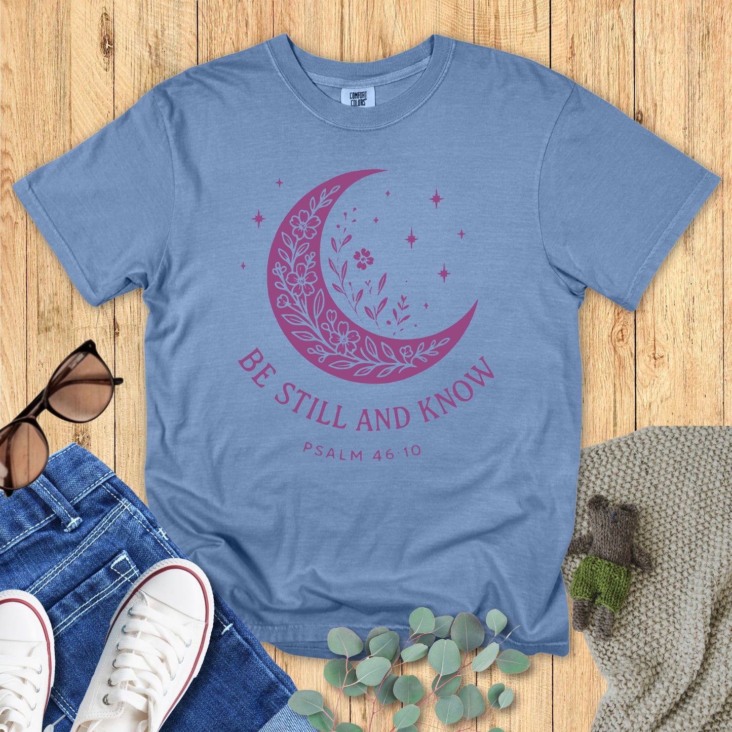 Blue t-shirt with pink crescent moon design and text on a wooden surface with accessories.