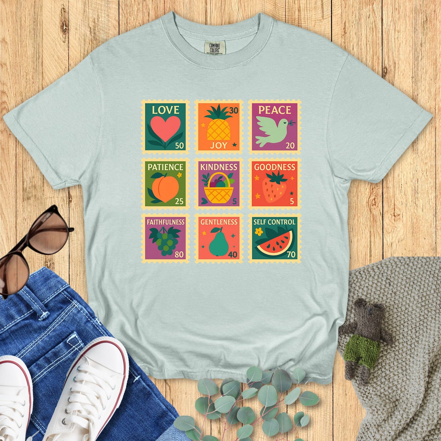Light blue t-shirt with colorful graphic design on a wooden surface with jeans and sneakers.