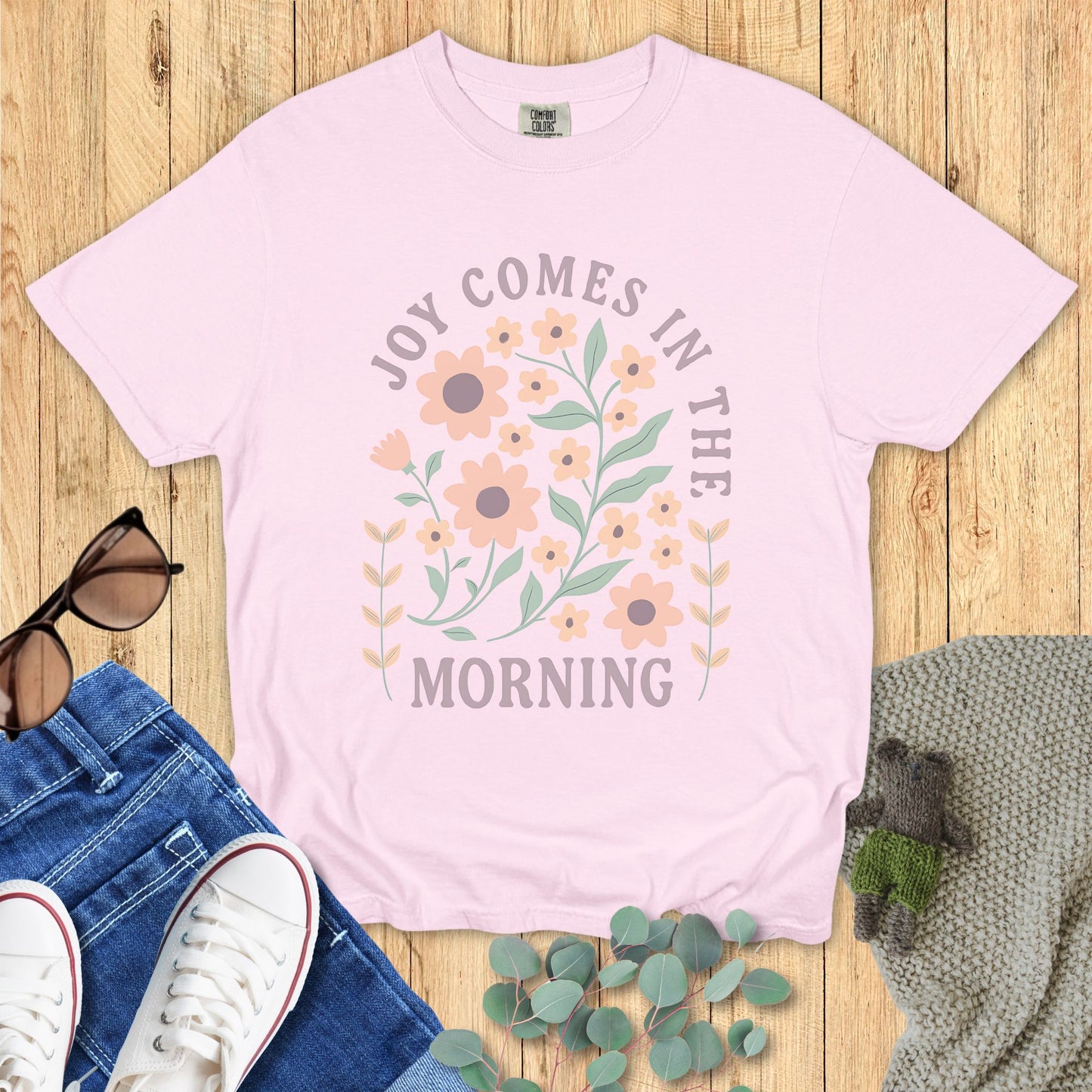 Pink t-shirt with floral design and text on a wooden surface with jeans and sneakers.