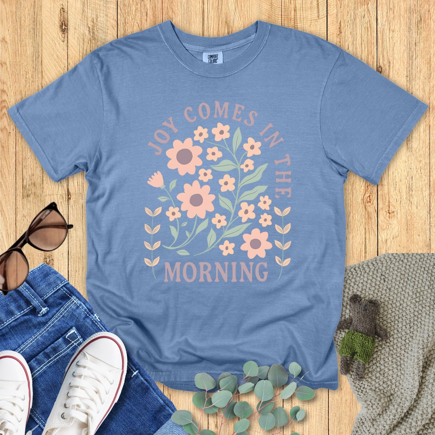 Blue t-shirt with floral design and text on a wooden surface with accessories.